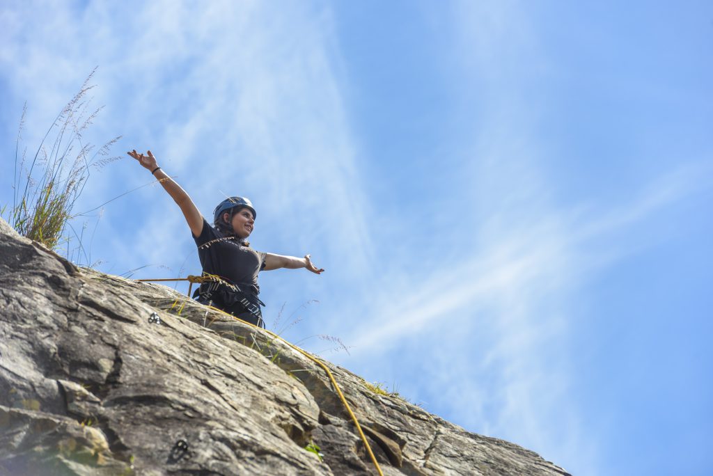 A young person in a climbing helmet and harness standing on a cliff with arms raised against a blue sky